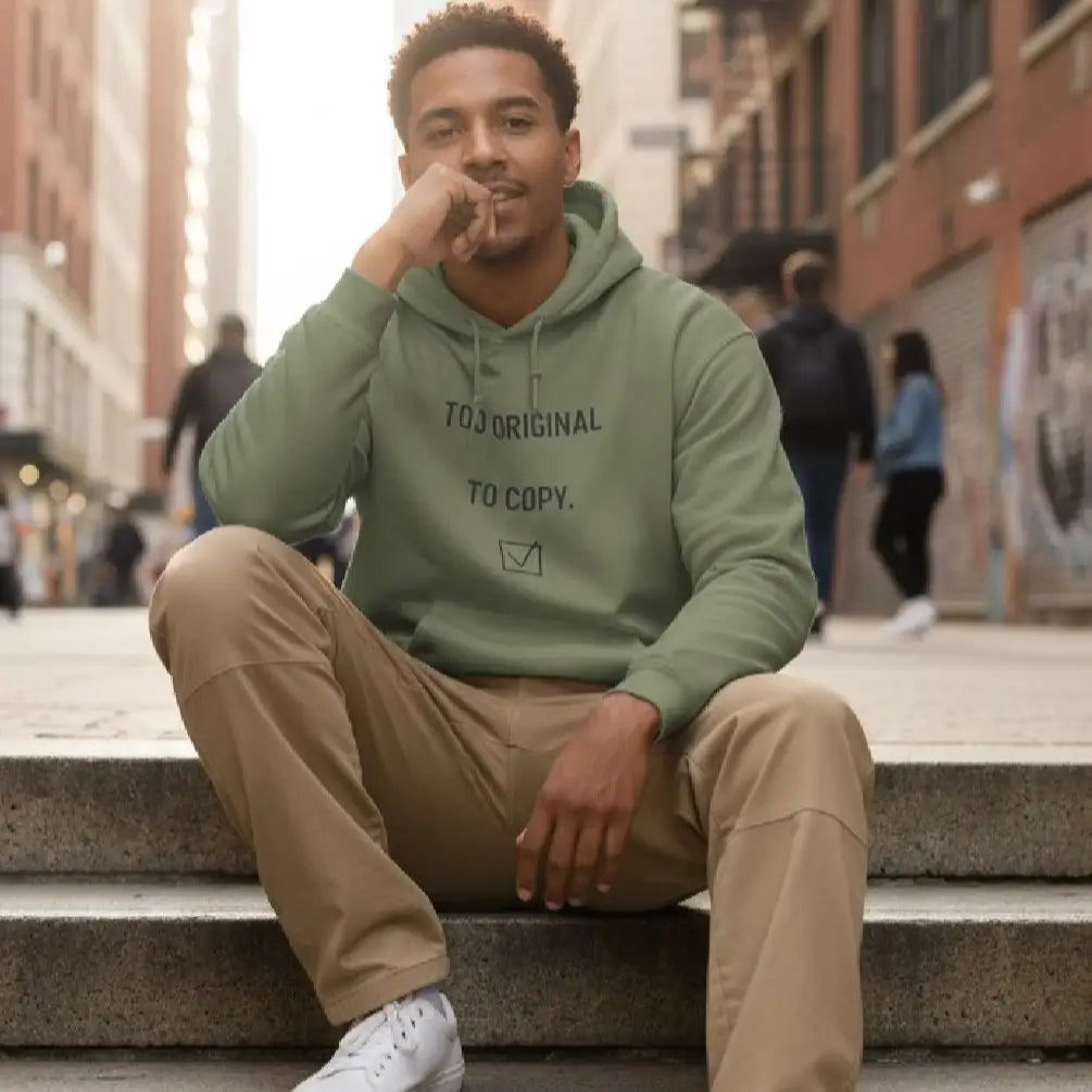 A man sitting on outdoor concrete steps in an urban setting, wearing a sage green hoodie with the text 'TO ORIGINAL TO COPY.' and khaki pants.